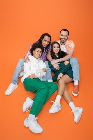 Hochwinkelaufnahme glücklicher und modischer multiethnischer Freunde auf orangefarbenem Hintergrund