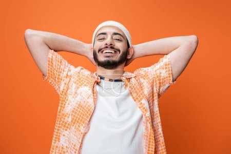 Foto de Hombre barbudo alegre en traje de moda posando con las manos detrás de la cabeza aislado en naranja - Imagen libre de derechos