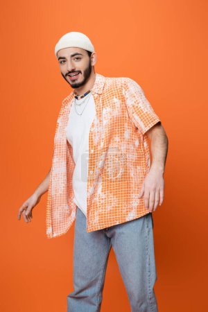 young bearded man in trendy casual attire smiling at camera while standing isolated on orange