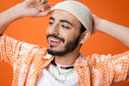 portrait of cheerful bearded man in beads touching white beanie and looking away isolated on orange