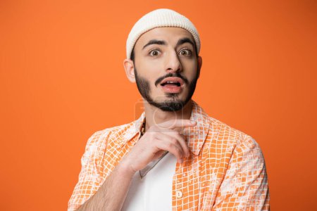impressed bearded man in trendy shirt and white beanie looking at camera isolated on orange