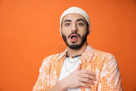 astonished bearded man in white beanie looking at camera isolated on orange