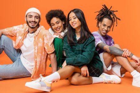 happy multiethnic friends in trendy attire sitting and smiling at camera on orange background