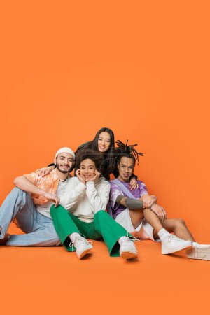 multiethnische Freunde in trendigen Klamotten sitzen und lächeln in die Kamera auf orangefarbenem Hintergrund