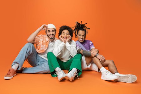 Photo for Joyful multicultural friends in stylish outfit sitting and smiling at camera on orange background - Royalty Free Image