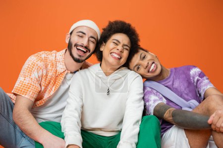 cheerful multiracial man looking at camera near friends laughing with closed eyes isolated on orange
