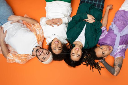 top view of joyful and stylish multicultural friends lying on orange background