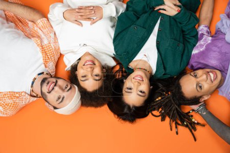 top view of pleased and trendy multiethnic friends lying and smiling at camera on orange background