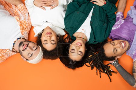 top view of smiling multiethnic friends in trendy clothes lying with closed eyes on orange background