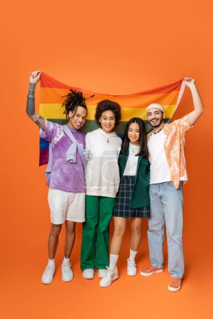 Foto de Longitud completa de sonrientes amigos multiétnicos en ropa de moda con bandera lgbt en naranja - Imagen libre de derechos
