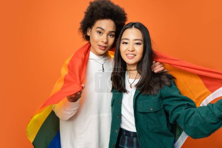 young multiracial women holding lgbt flag and smiling at camera isolated on orange