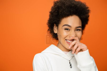Téléchargez les photos : Portrait de femme afro-américaine gaie tenant la main près du visage et regardant la caméra isolée sur orange - en image libre de droit