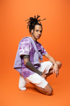 full length of multiracial man in trendy outfit sitting on haunches and looking at camera on orange background