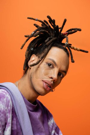 Foto de Portrait of fashionable multiracial man with piercing and dreadlocks looking at camera isolated on orange - Imagen libre de derechos