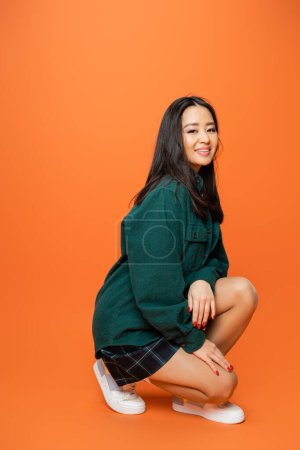full length of happy asian woman in trendy outfit posing on haunches on orange background
