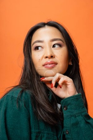Porträt einer brünetten asiatischen Frau mit Make-up und roter Maniküre, die isoliert auf Orange wegschaut