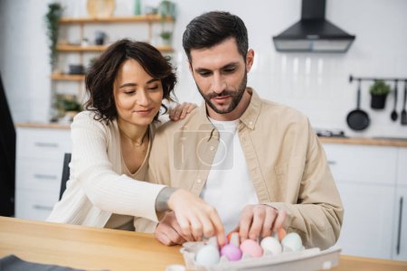 Smiling woman and man putting Easter eggs in tray at home 