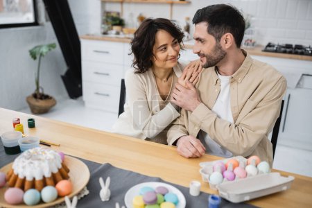 Smiling couple looking at each other near Easter eggs and cake at home
