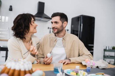 Smiling woman holding paintbrush near husband with Easter egg in kitchen 
