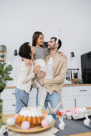Parents holding daughter near blurred Easter cake and eggs at home 