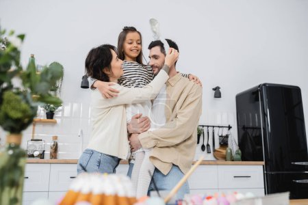 Smiling girl hugging parents near blurred Easter eggs in kitchen 