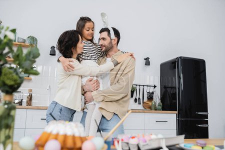 Positive parents hugging daughter near blurred Easter eggs and cake in kitchen 