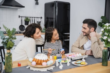 Smiling woman and daughter looking at man near Easter eggs and cake at home 