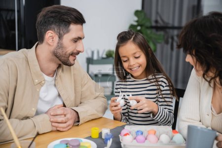 Smiling girl holding Easter rabbits near cheerful parents and food at home 