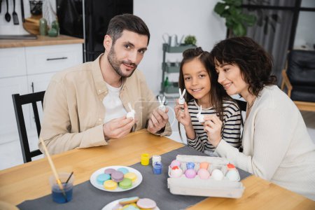 Parents and child holding Easter decor near eggs and macaroons in kitchen 