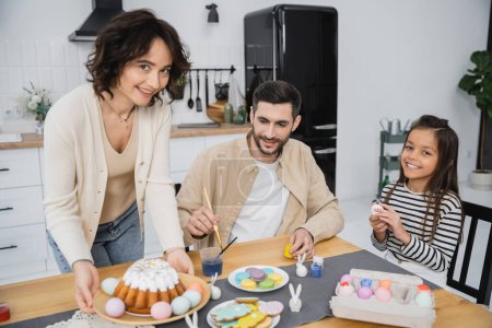 Family coloring Easter eggs near cheerful mother holding plate with cake at home 