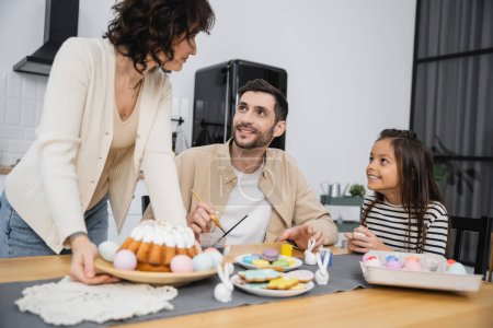 Familia sonriente huevos para colorear cerca de la mujer ronroneo plato con pastel de Pascua en la mesa en casa 