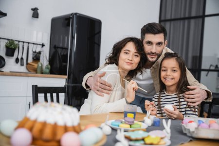 Man hugging family with paintbrushes near Easter eggs and cake at home 