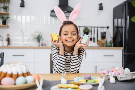 Fröhliches Kind mit Stirnband hält Plätzchen neben Osterkuchen und Eiern zu Hause 