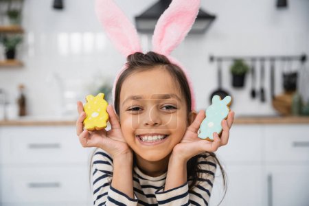 Smiling kid in Easter headband with bunny ears holding cookies in kitchen 