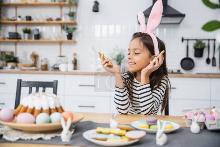 Lächelndes Kind mit Stirnband betrachtet Osterkekse in der Nähe von Lebensmitteln in der Küche 