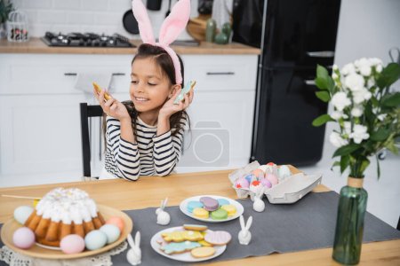 Happy preteen kid in headband holding Easter cookies near flowers and food at home 