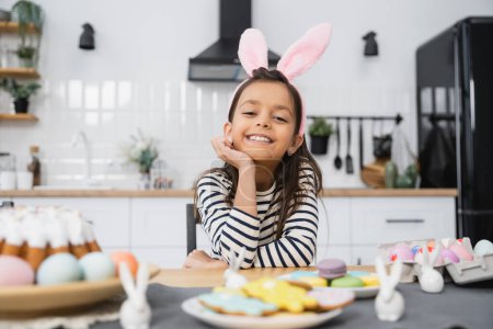 Fröhliches Mädchen mit Hasenohren-Stirnband sitzt neben Osterkuchen und Eiern zu Hause 