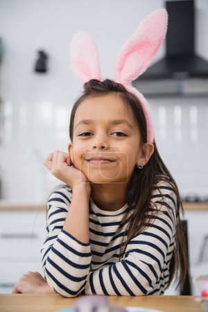 Smiling girl in Easter bunny ears headband looking at camera at home 