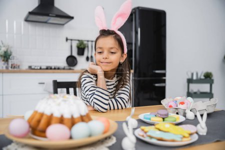 Preteen kid in bunny ears headband looking at Easter cake and eggs in kitchen 