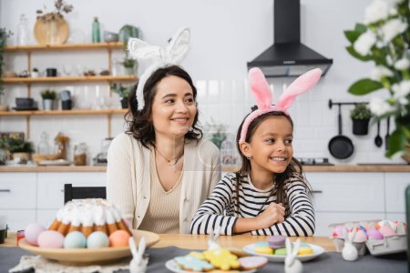 Smiling mother and kid in bunny ears headbands sitting near Easter cake and eggs at home 