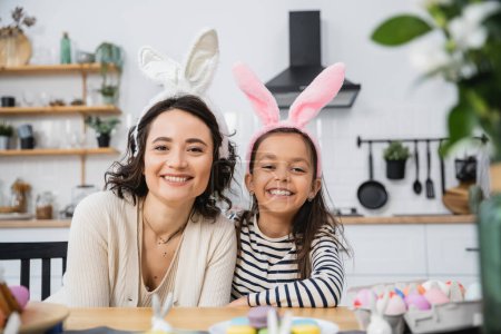 Sonriente mamá y el niño en cintas de Pascua mirando a la cámara cerca de los huevos y macarrones en casa 