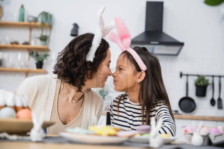 Mother and kid in Easter headbands sitting nose to nose in kitchen 