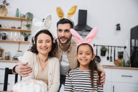 Smiling man hugging wife and daughter in Easter headbands in kitchen 