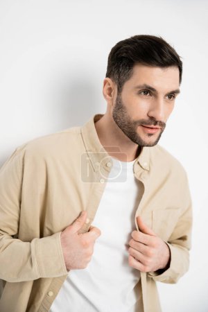 Foto de Hombre barbudo en camiseta tocando camisa beige sobre fondo blanco - Imagen libre de derechos