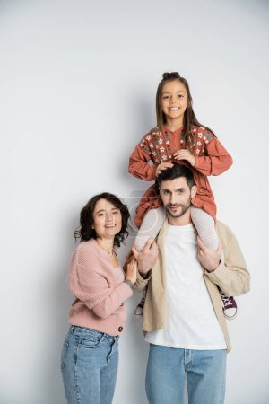 Man holding cheerful daughter on shoulders near wife on white background 