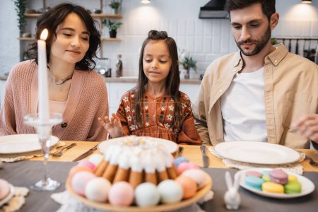 Family sitting near blurred Easter cake and candle on table at home 