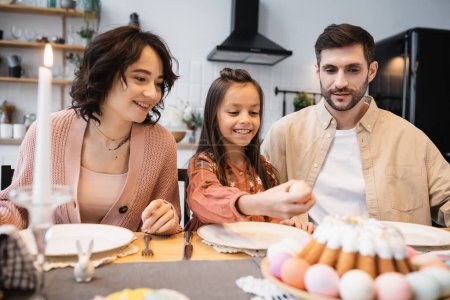 Smiling parents looking at child holding Easter egg during festive dinner at home 
