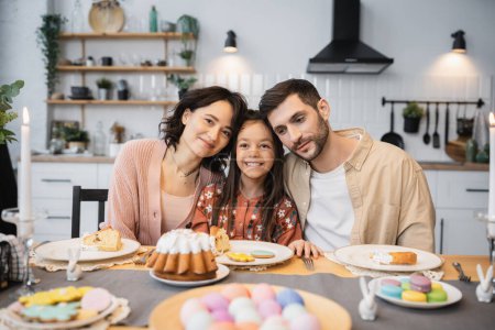 Cheerful parents and kid sitting near festive Easter dinner in kitchen 