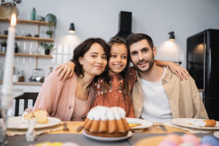 Preteen girl hugging parents near Easter cake and candle at home 
