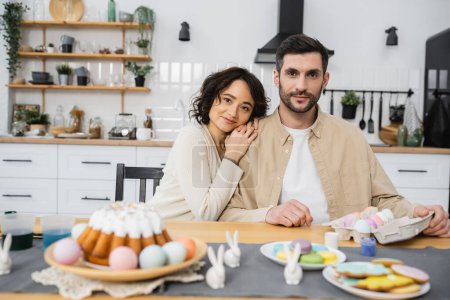 Smiling woman hugging husband near paints and Easter food at home 
